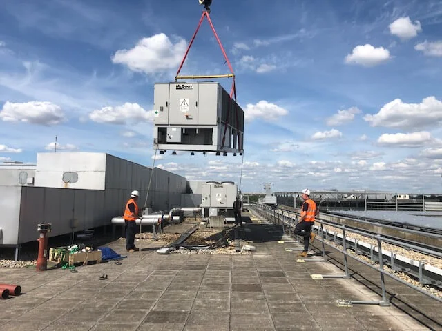 Two workers in safety gear supervise a large industrial unit being lifted by a crane onto a rooftop under a blue sky with scattered clouds. Tools and equipment are visible on the roof.