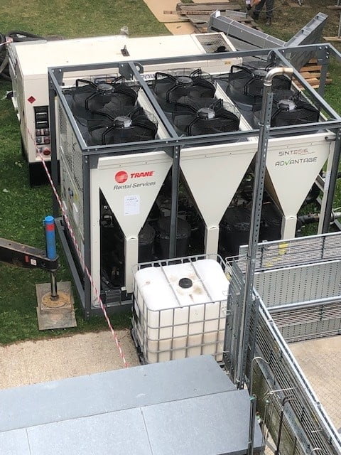 A large industrial air conditioning unit labeled Trane Rental Services is set up outdoors, next to a white water tank and surrounded by safety barriers and equipment.
